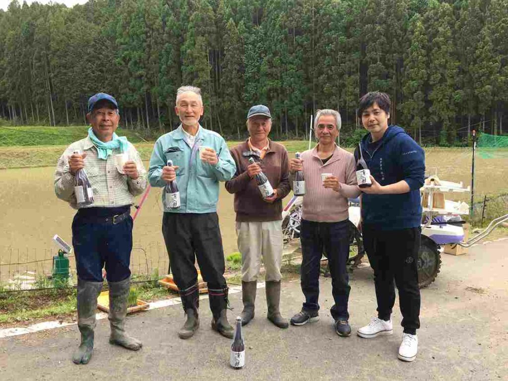 équipe de la brasserie matsui shuzo devant un champ de riz, tenant des bouteilles de saké